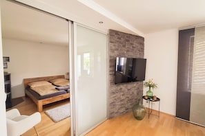 A modern bedroom with a bed, a TV mounted on a stone wall, a small table with a vase, and a sliding door leading to another room.