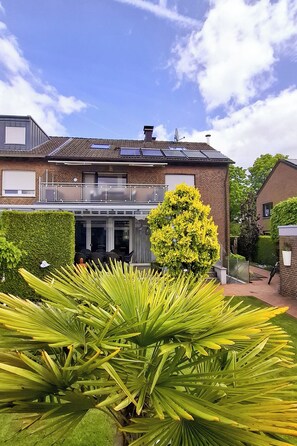 Une maison à deux étages dotée d’un balcon, de panneaux solaires et d’un jardin bien entretenu avec un grand palmier.