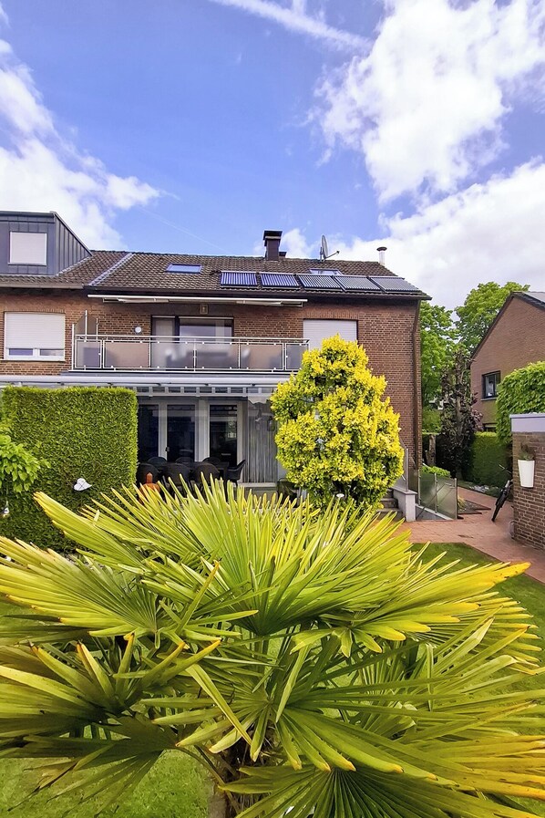 A two-story house with a balcony, solar panels, and a well-maintained garden with large green plants.