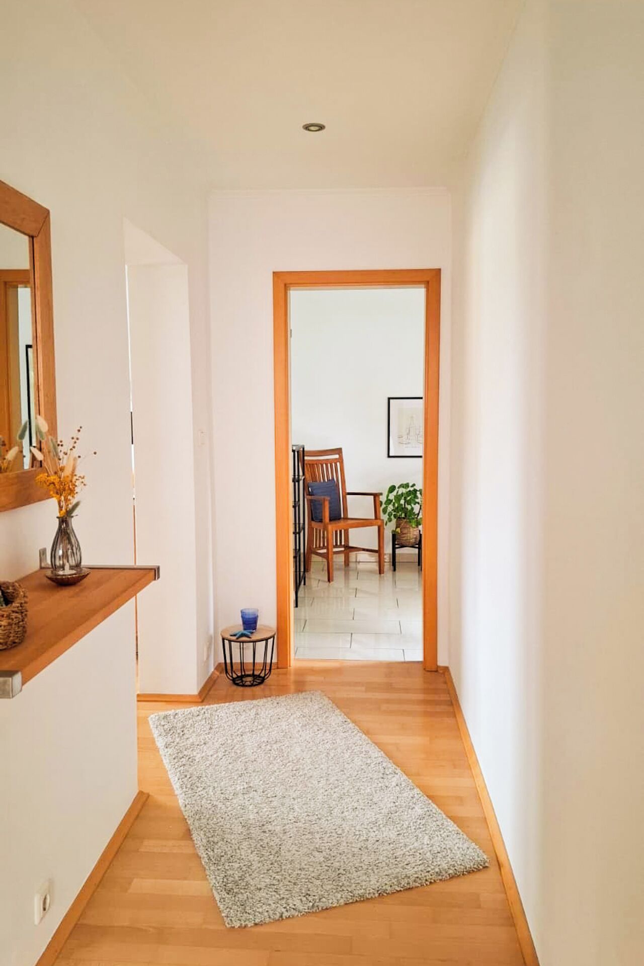 A hallway with a wooden floor, a white rug, a chair, a table, and a plant.
