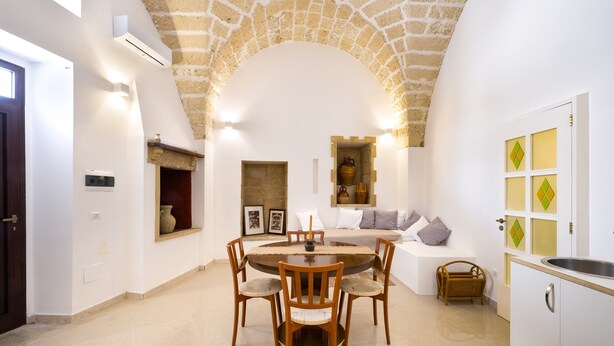 A dining area with a round table and chairs, a stone arch ceiling, a kitchen with a sink, and a built-in bench with cushions. - Apartment in 'Dimora S' Hotel with Private Terrace, Wi-Fi, and Air Conditioning (Sogliano Cavour)