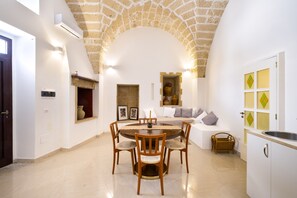Un espace repas avec une table ronde et des chaises, un plafond en forme d’arc en pierre, une cuisine avec un évier et un banc intégré agrémenté de coussins.