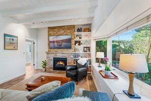 A living room with a fireplace, a television, a bookshelf, and a window with a view of greenery. - Casa Alma del Mar | Hot Tub + Walk to Beach (Santa Barbara)