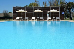 Une piscine entourée de chaises longues blanches et d’ombrelles, une clôture en bois et un panneau indiquant un complexe hôtelier en bord de mer.