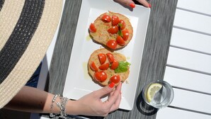 Una persona sostiene un plato con dos sándwiches abiertos cubiertos de tomate y albahaca, un vaso de agua de limón y un tapete tejido.