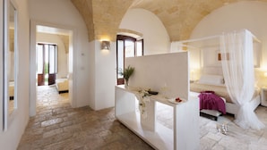 A bedroom with a stone floor, a bed with a canopy, a bedside table with a vase, and a view of a stone archway.