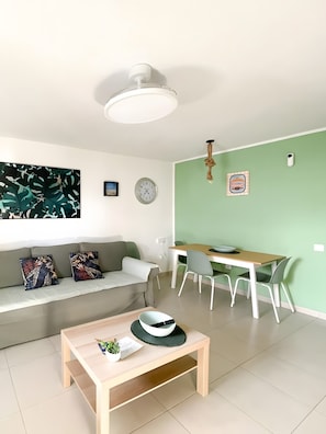 Een moderne woonkamer met een bank, een eettafel met stoelen, een plafondlamp, een wandklok en een ingelijst kunstwerk.