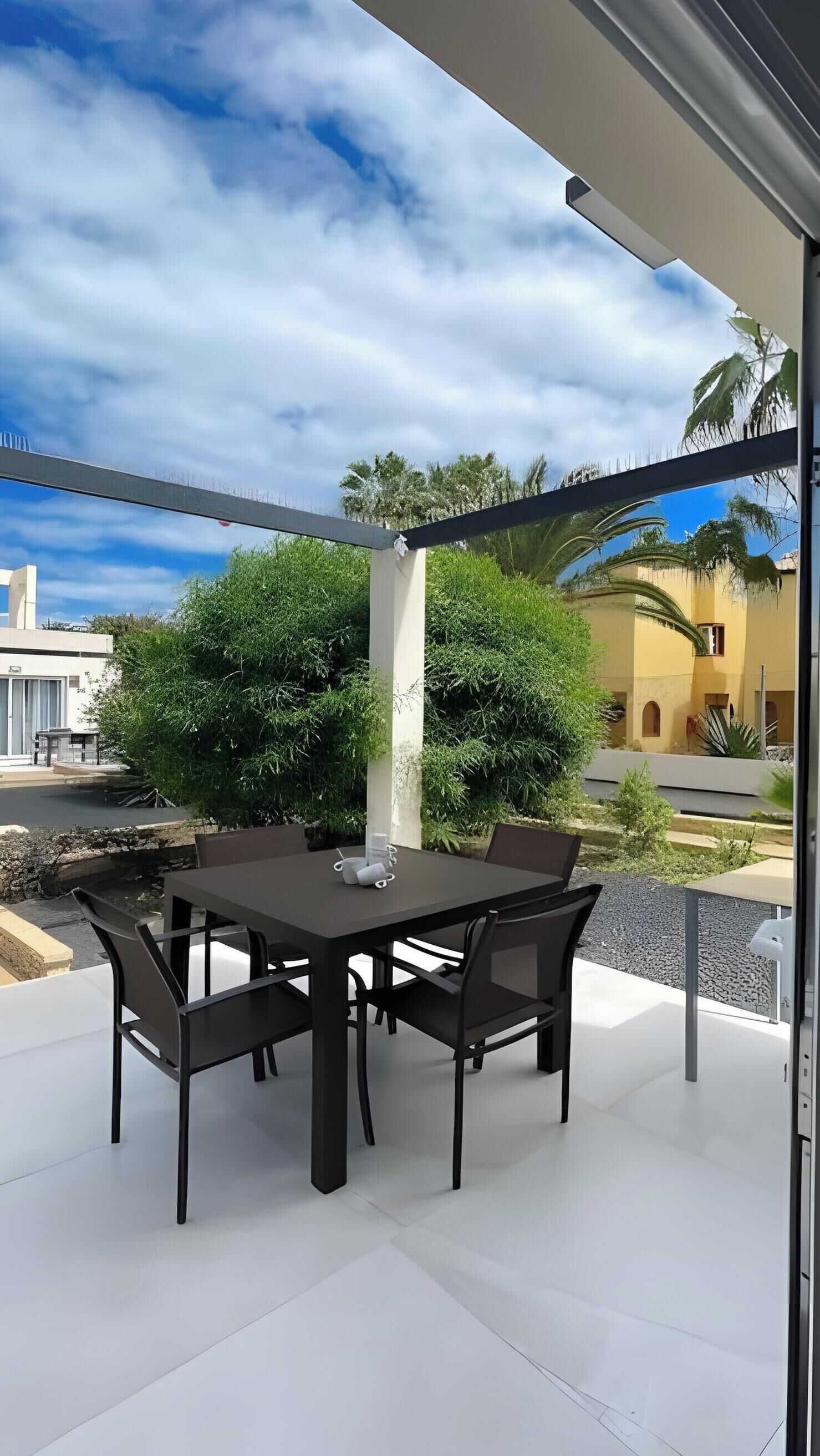 Un patio al aire libre con una mesa y sillas, vista a un edificio y cielo despejado.