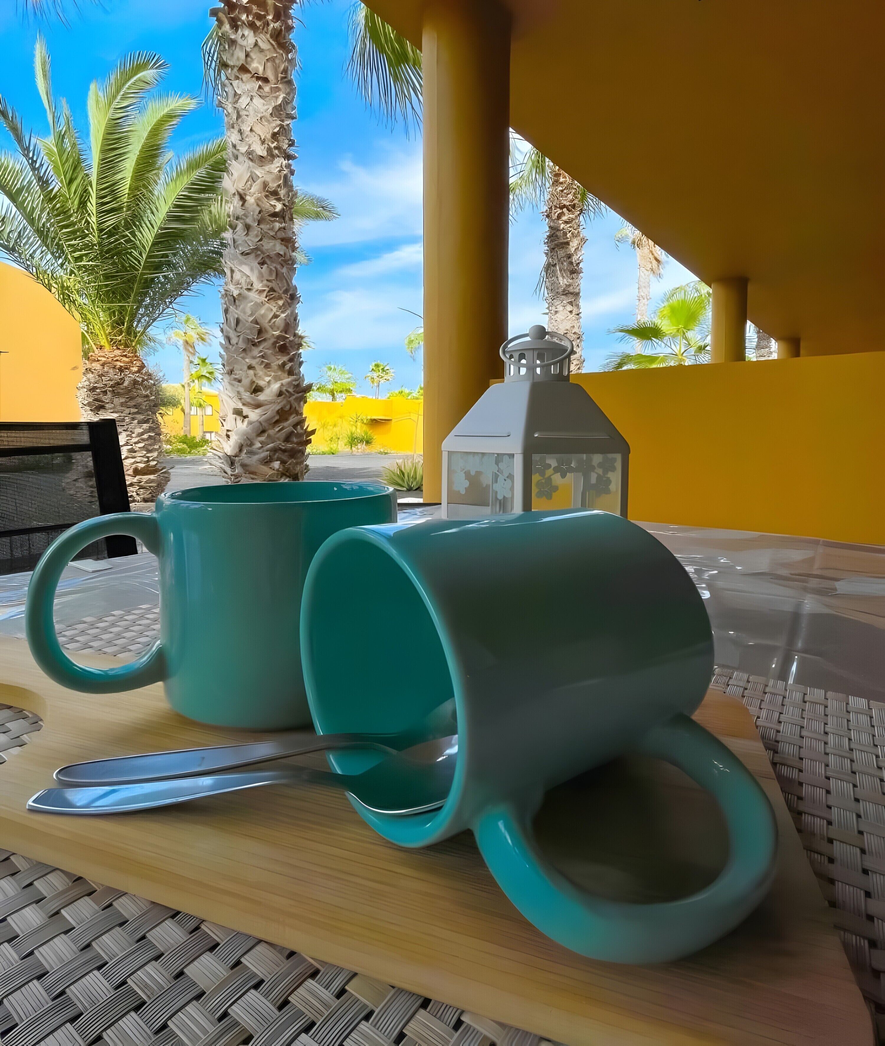 Two teal mugs on a wooden table with a woven mat underneath, a metal spoon, and a white lantern in a yellow-walled outdoor area with palm trees.