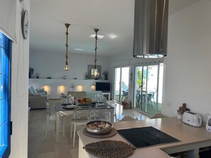 A modern kitchen with a dining area, a large window, and a TV in the background. - Bahiazul Villa Opale by B-Rent Corralejo (La Oliva)