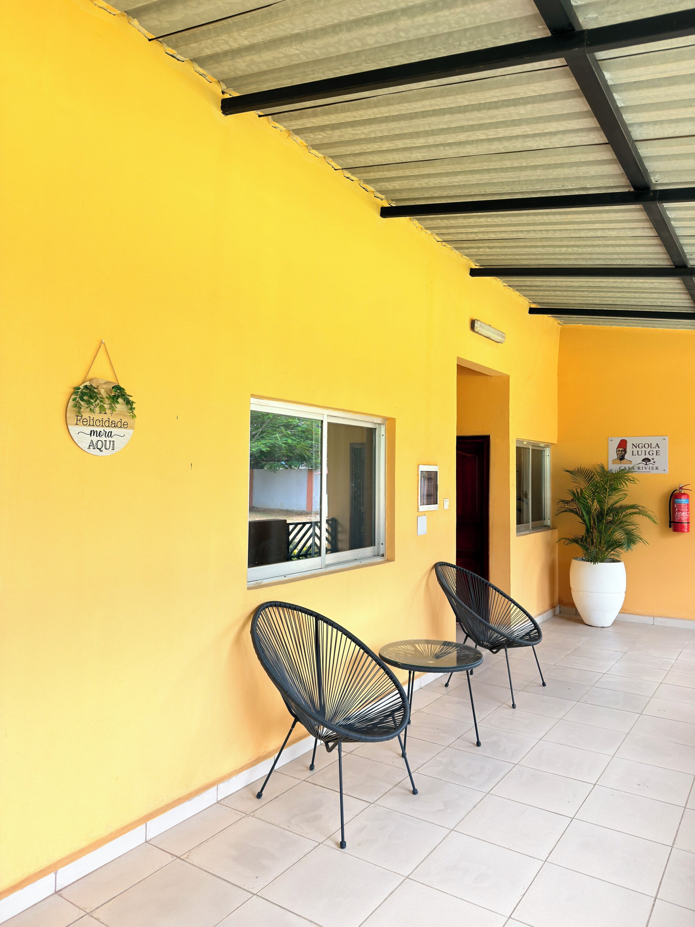 A yellow exterior wall with a metal roof, a window, two black chairs, a small table, a potted plant, and a sign with a name.