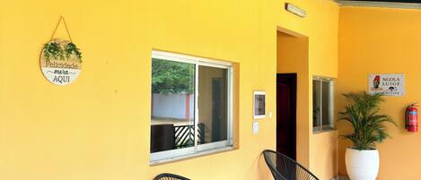 A yellow exterior wall with a metal roof, a window, two black chairs, a small table, a potted plant, and a sign with a name.
