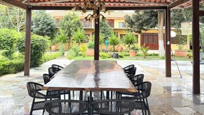 A covered outdoor dining area with a long table and chairs, surrounded by greenery and a building in the background.