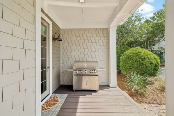 Un espace extérieur abrité comprenant un barbecue, une porte donnant accès à un garage et un petit jardin agrémenté d’un arbuste et d’une plante.
