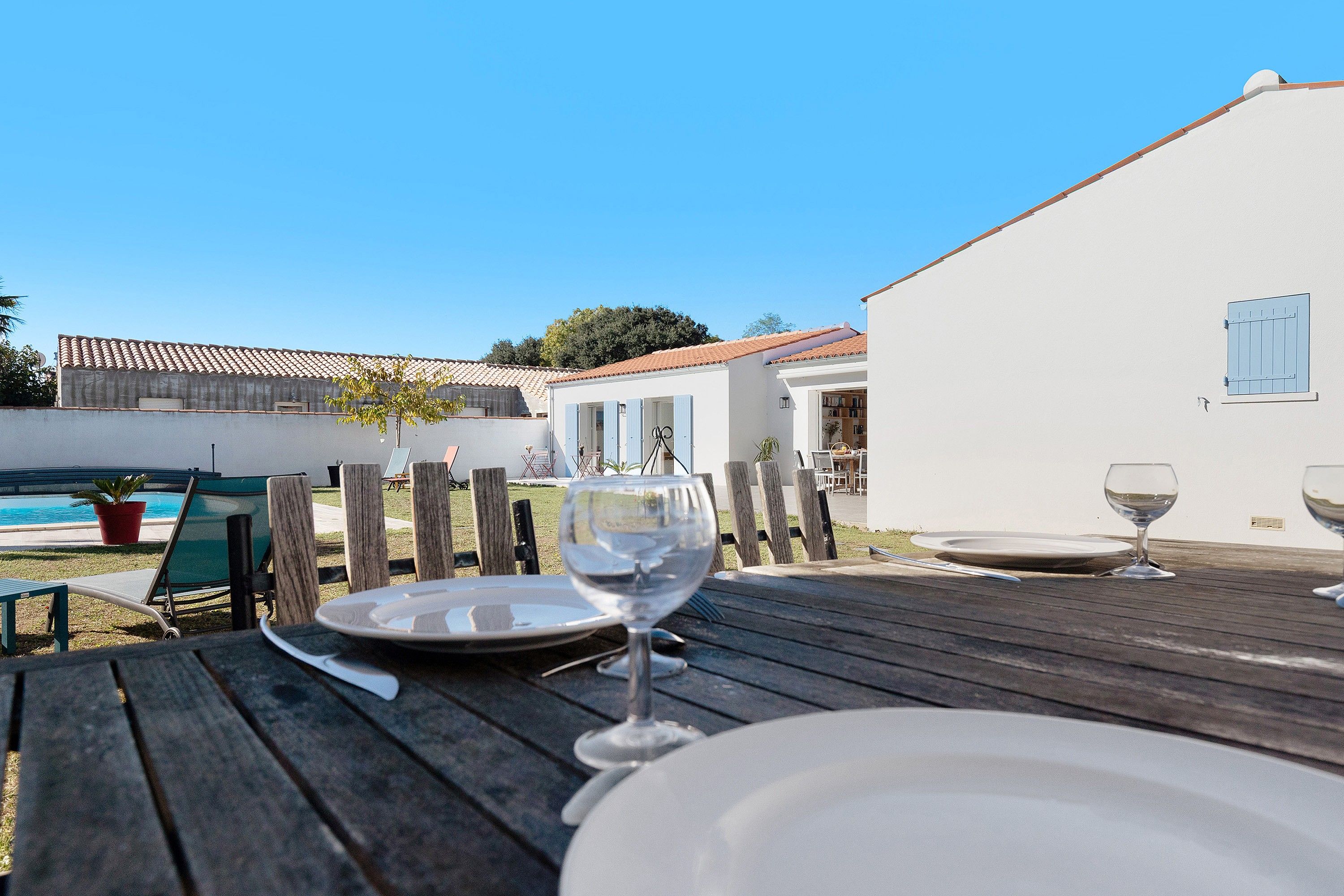 A wooden deck with a table set for dining, overlooking a swimming pool and a white building with blue shutters.