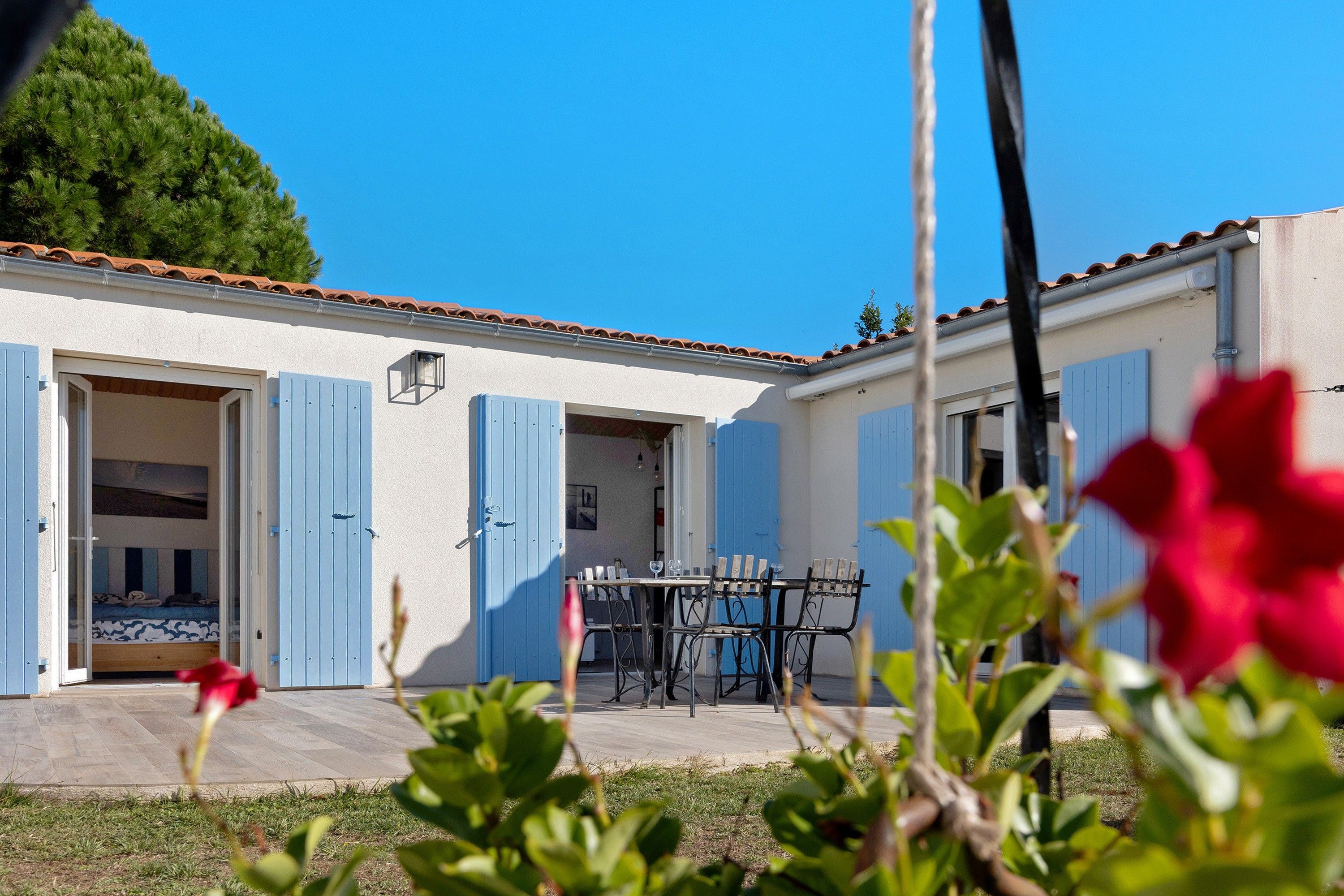 Un bâtiment à deux étages avec des volets bleus, un patio comprenant une table et des chaises, et un ciel d’un bleu éclatant.