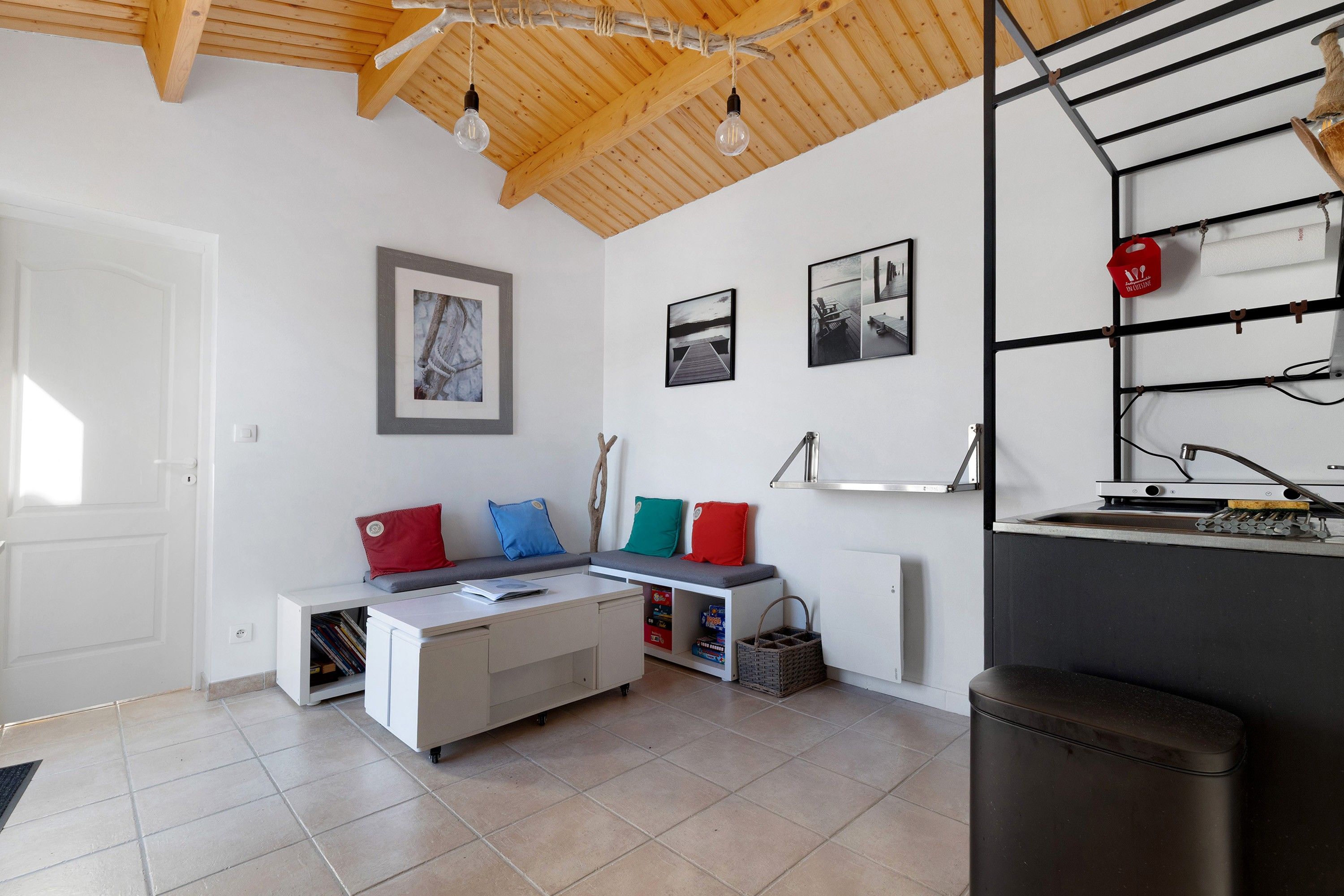 Een moderne woonkamer met een witte bank, een klein tafeltje en een boekenkast. Er hangen afbeeldingen in lijsten aan de muur en het plafond is van hout.
