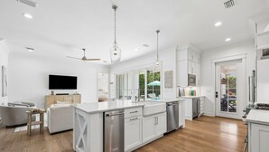A modern kitchen with white cabinetry, a central island, and a dining area with a sofa and table. - Retreat Close to Beachw/ Pool and Screened Porch (Santa Rosa Beach)