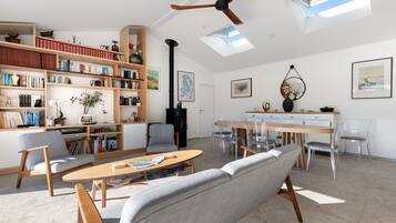 Un vaste salon doté d’un haut plafond, d’un ventilateur de plafond, d’une cheminée, d’un coin repas avec une table et des chaises en bois, et d’une grande bibliothèque remplie de livres.