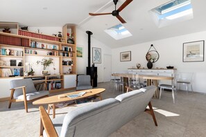 A spacious living room with a high ceiling, a ceiling fan, a fireplace, a dining area with a wooden table and chairs, and a large bookshelf filled with books. - Bed and Breakfast 'Suite Les Cabanes' with Shared Pool, Shared Garden and Wi-Fi (La Brée-les-Bains)