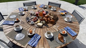 Une table ronde en bois, dressée à l’extérieur et entourée de chaises, sur laquelle sont disposés divers aliments pour le petit-déjeuner, notamment des viennoiseries, du pain et des boissons.