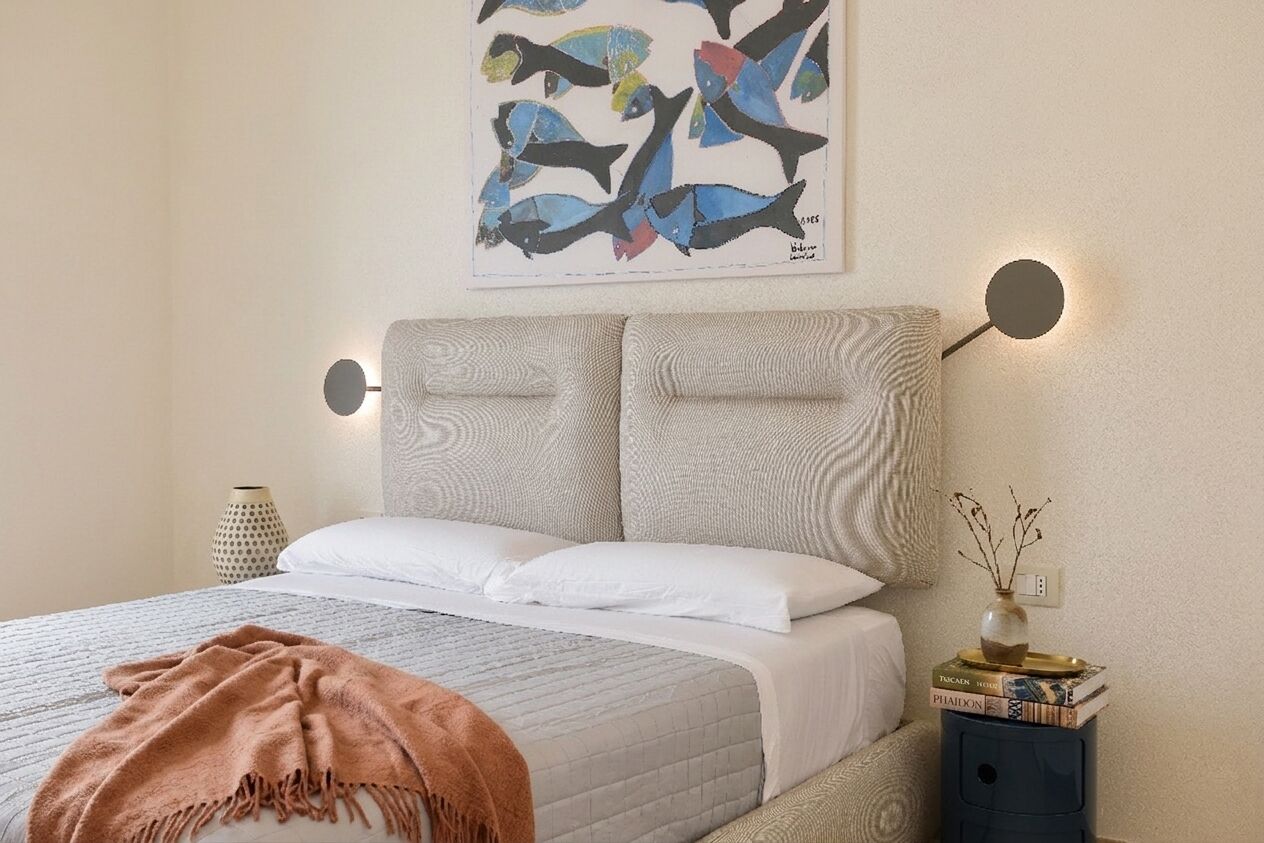 Una cama con un cabecero texturizado, una mesita de noche con un florero y libros, y una obra de arte colgada en la pared.