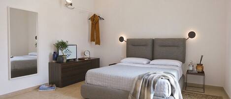 A bedroom with a bed, bedside table, nightstand, mirror, and wall-mounted lamp.