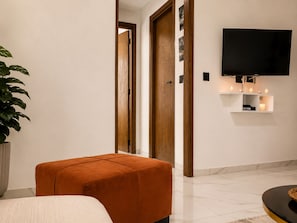 A modern interior with a wooden door, a flat-screen TV mounted on the wall, a potted plant, and a seating area with an orange ottoman. - Sandra 28 (Tunis)