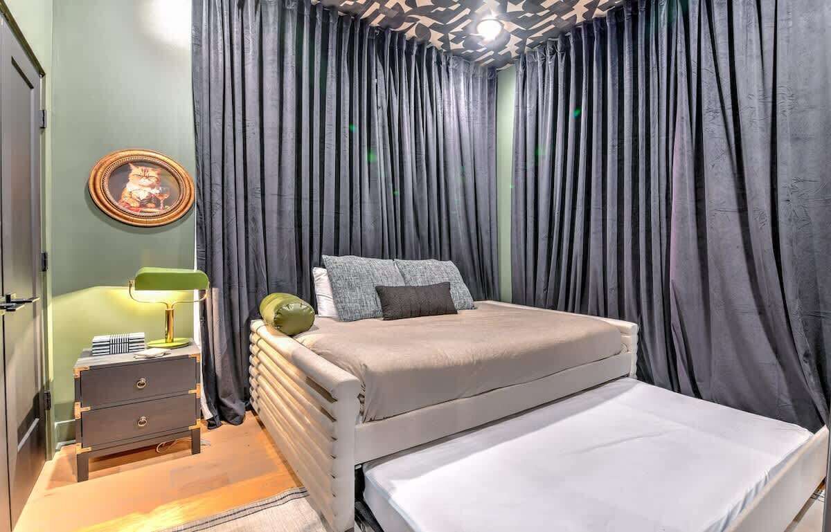 Een slaapkamer met een bed, een nachtkastje met een lamp, een lijst met een afbeelding aan de muur, en donkere gordijnen.