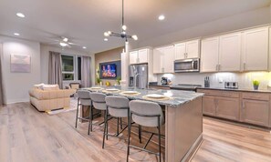 A modern kitchen with a central island, bar stools, a dining area with a sofa, and a TV in the background. - Lux Nash Experience | Firepit + Game Room. 9 Beds! (Nashville)