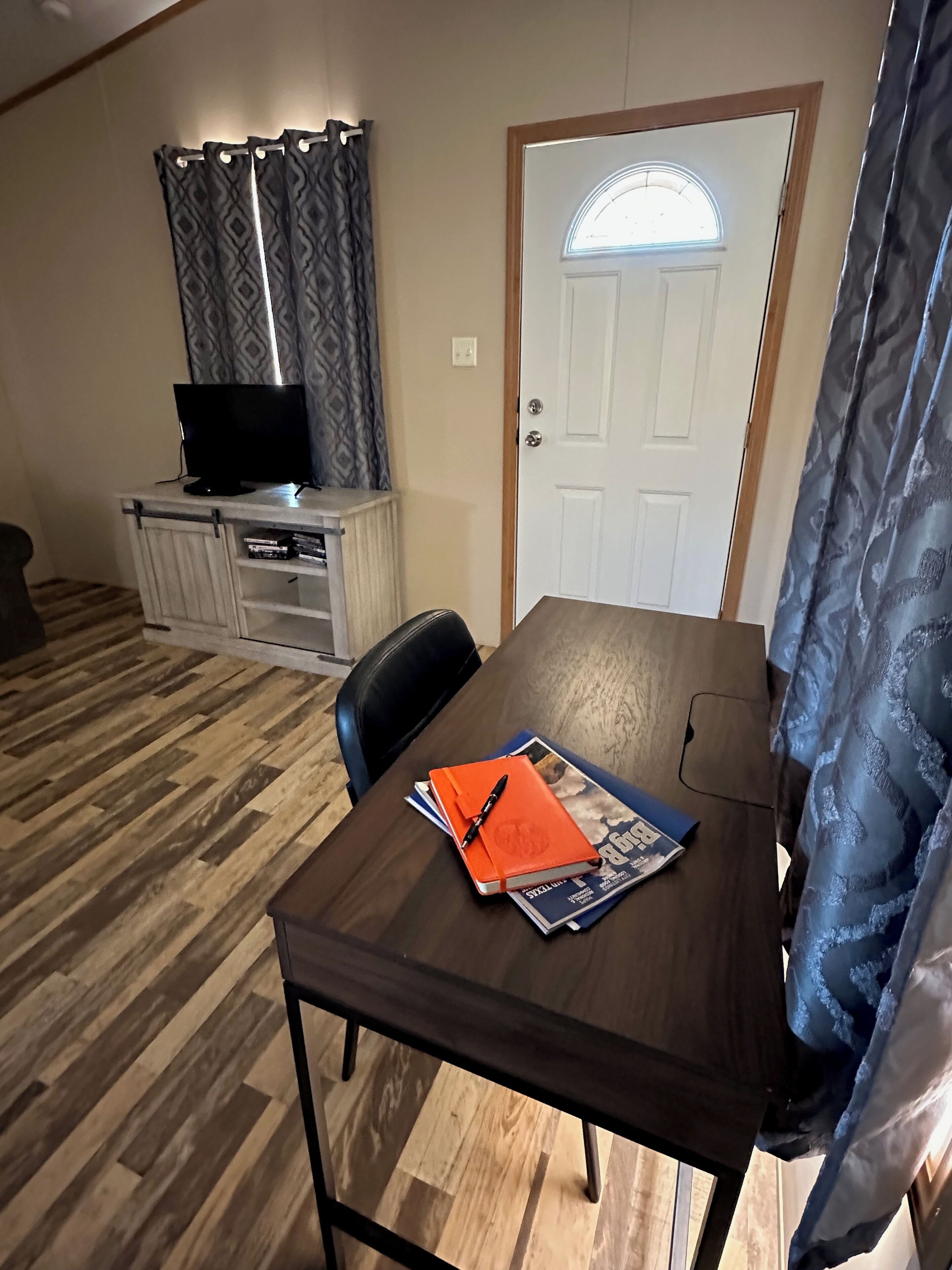 Una habitación con piso de madera, una mesa con un cuaderno y bolígrafo, un televisor sobre un soporte y una puerta con ventanal.
