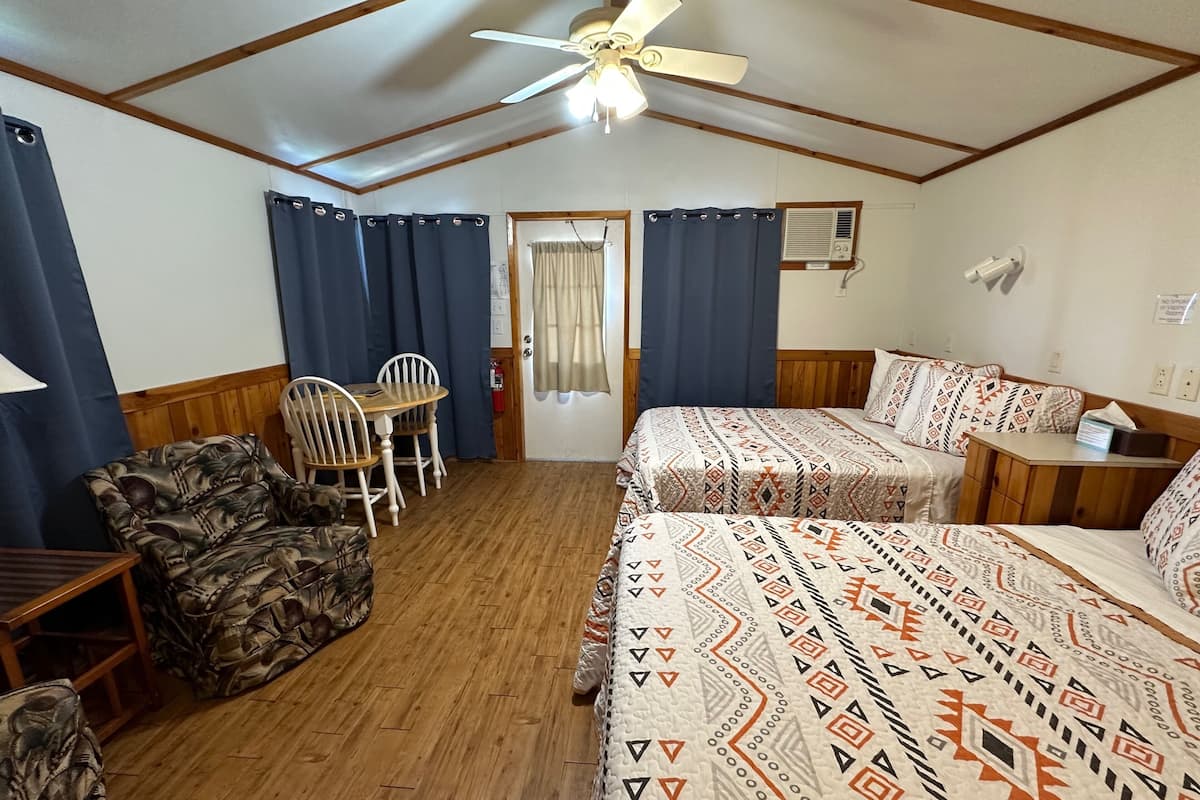 Una habitación con ventilador de techo, una mesa de comedor de madera con sillas, dos camas con ropa de cama estampada y una ventana con cortinas.