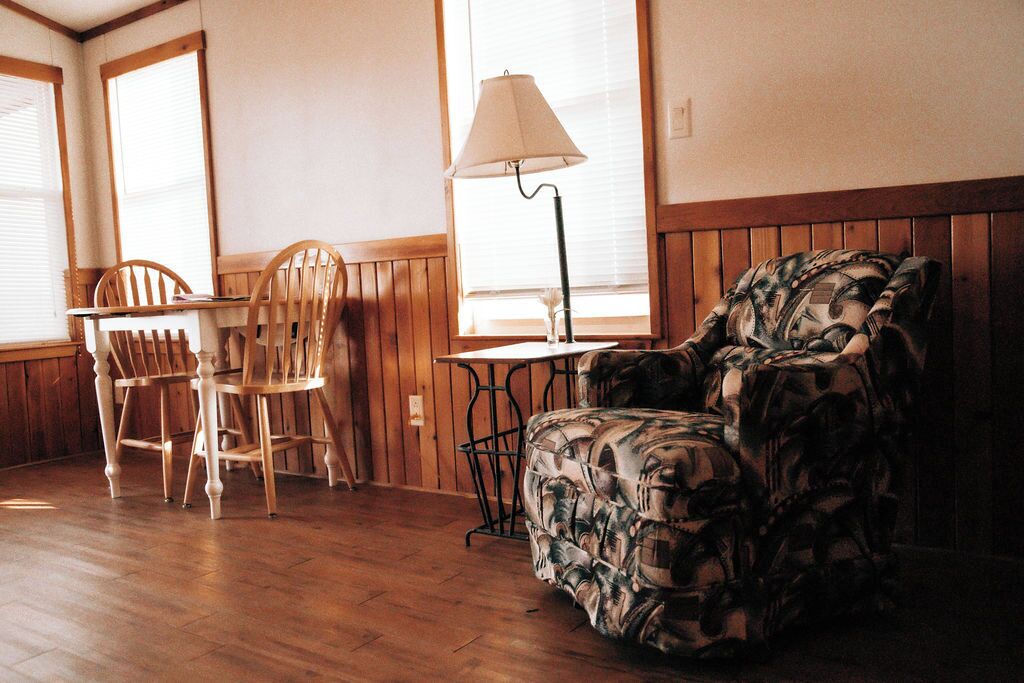 Una habitación con suelo de madera, una mesa con sillas, un sillón estampado y una lámpara.