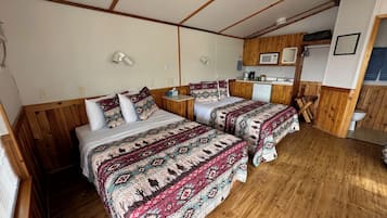 A room with two beds, wooden walls, a small kitchenette, and a ceiling fan.