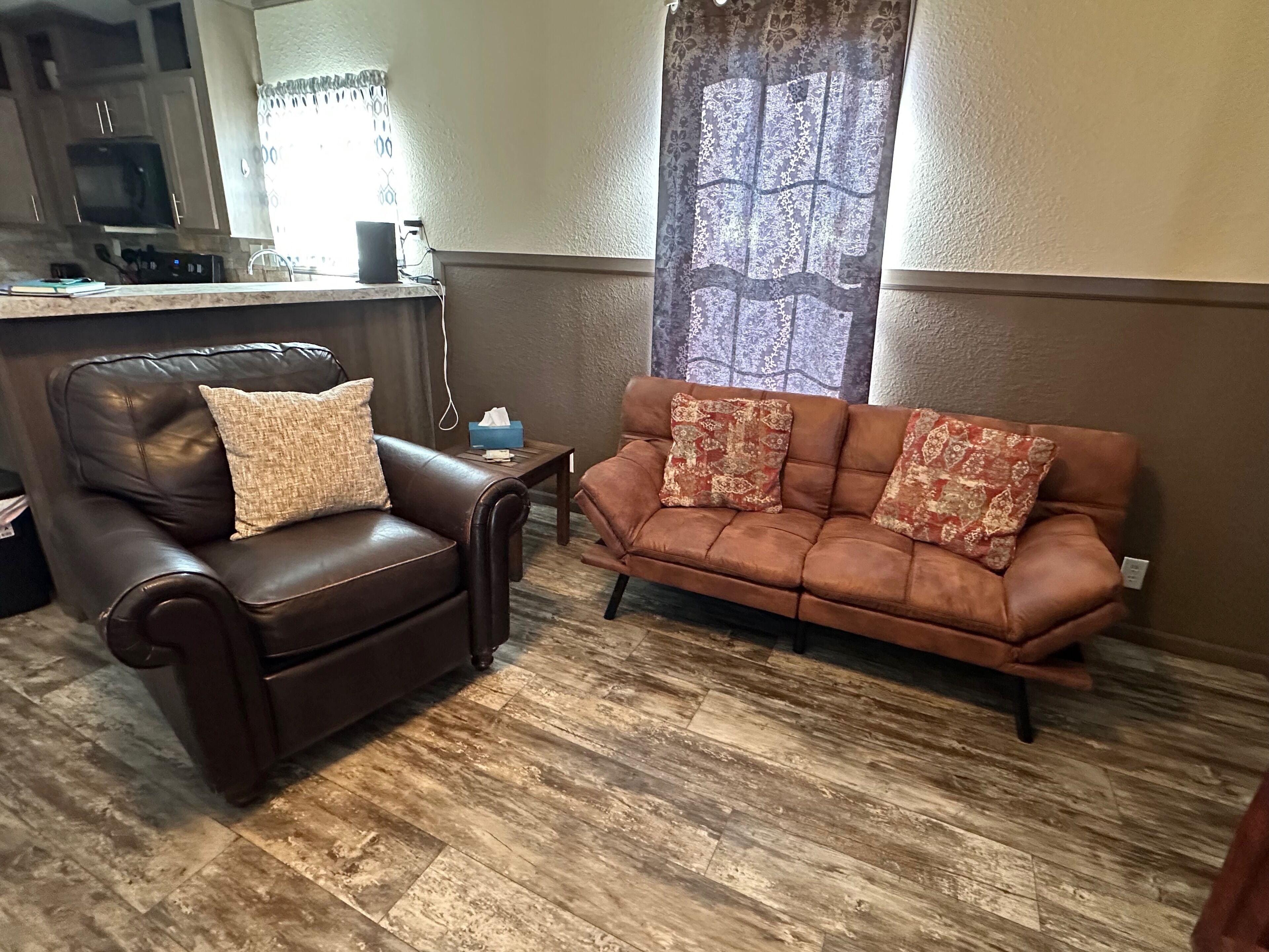 Una sala de estar con un sofá de cuero marrón, un sillón de cuero marrón y piso de madera.