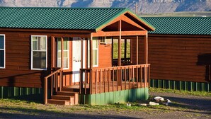 A wooden cabin with a green roof, a porch, and a view of mountains.