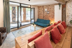 Ein modernes Wohnzimmer mit einem blauen Sofa, einer Holzbänkl mit roten Kissen, einem kleinen Tisch mit Pflanze und einer Ziegelwand.