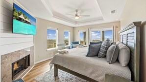 Une chambre avec une cheminée, un lit avec des oreillers, un ventilateur de plafond et une grande fenêtre donnant sur un beau paysage.