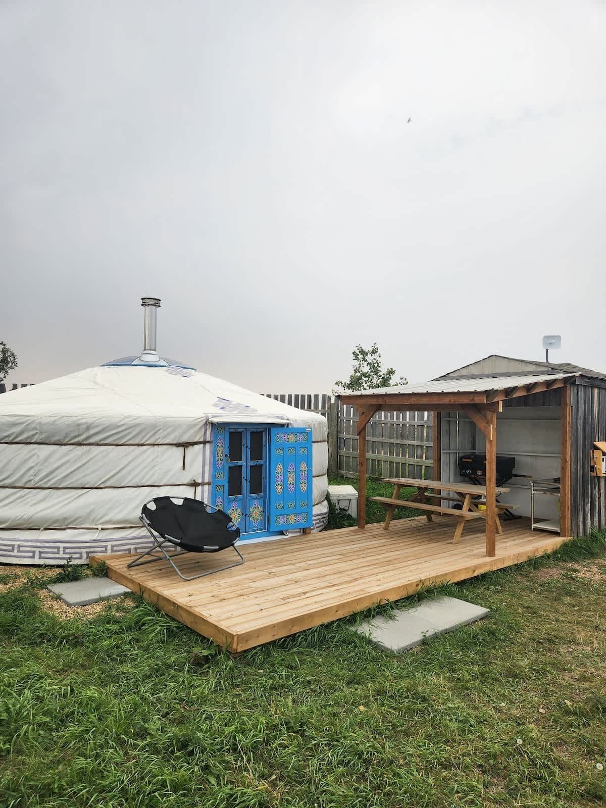 Una yurta con puerta azul, una silla negra y una terraza de madera con zona de barbacoa.