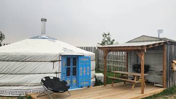 Una yurta con puerta azul, una silla negra y una terraza de madera con zona de barbacoa.