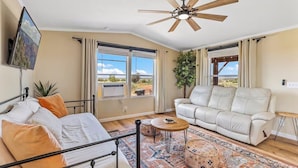 A room with a bed, two white leather sofas, a ceiling fan, a TV mounted on the wall, and large windows with curtains. - Stargazing Oasis | A/C | 35 Min to Grand Canyon (Williams)