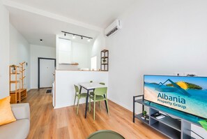 Una sala de estar moderna con una televisión mostrando 'Albania Property Group', un comedor con sillas, un sofá y piso de madera.