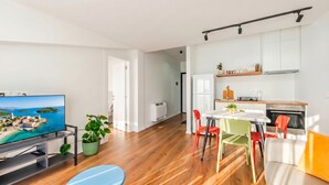 Ein modernes Wohnzimmer mit Flachbildfernseher, Holzboden, einer Couch, einem Esstisch mit bunten Stühlen und einer Küchenzeile mit Schränken und Geräten.