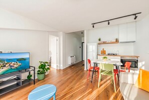 Una sala de estar moderna con una televisión de pantalla plana, piso de madera, un sofá, una mesa de comedor con sillas de colores, y una zona de cocina con gabinetes y electrodomésticos.