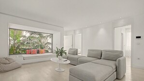 En moderne stue med en hjørnesofa, et lille hvidt bord og et vindue med udsigt til grøn bevoksning.