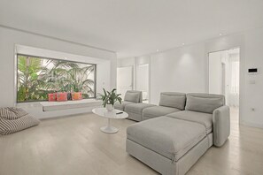 Ein modernes Wohnzimmer mit einer Eckcouch, einem kleinen weißen Tisch und einem Fenster mit Blick auf Grünflächen.