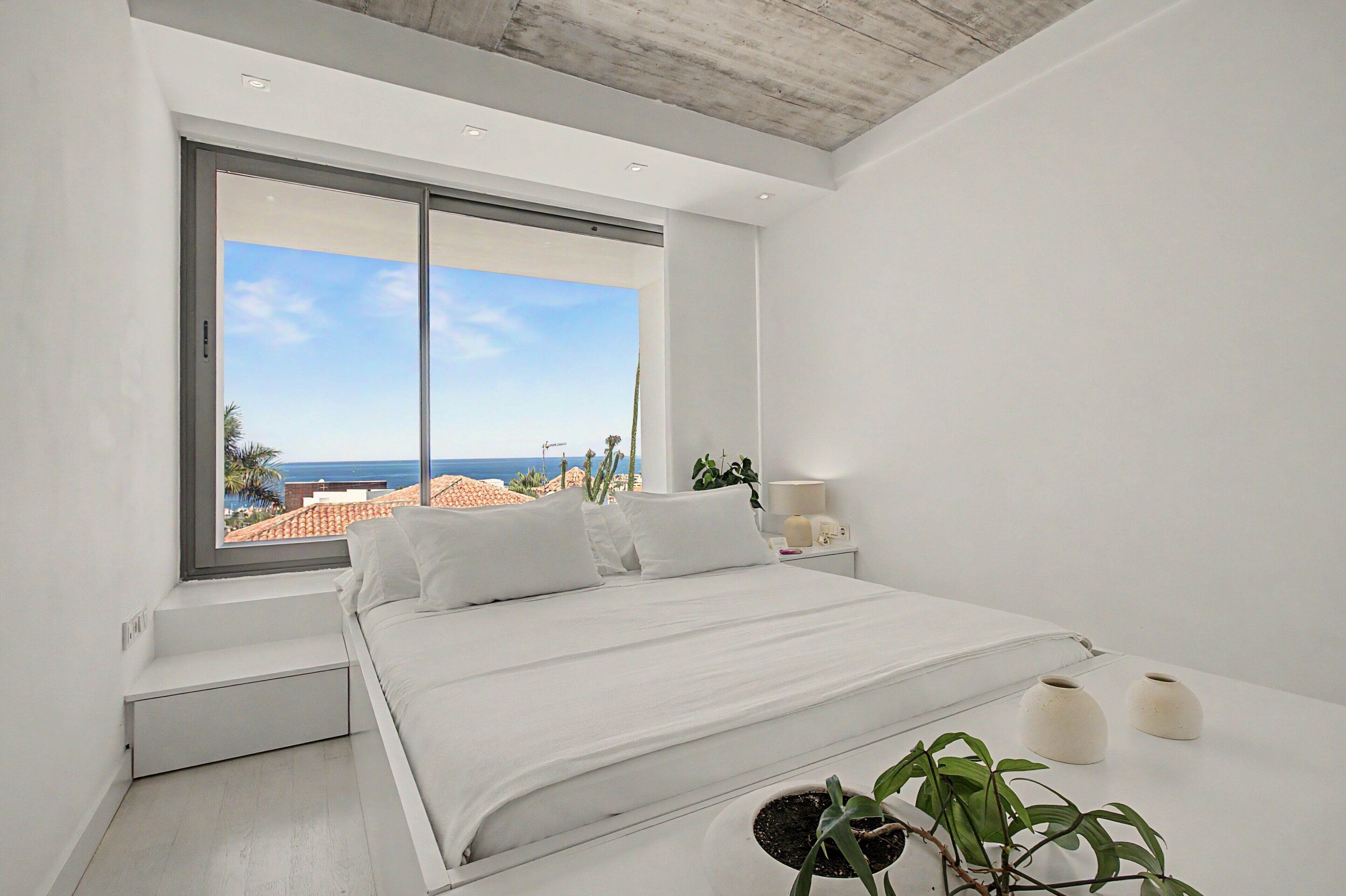 A modern bedroom with a large window, a bed, bedside tables, and a view of the sea.