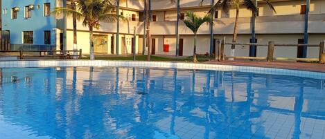 A swimming pool with tiled flooring, surrounded by palm trees and a multi-story building with balconies.