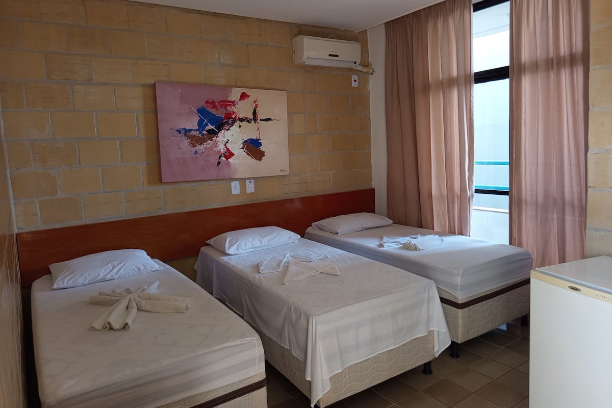 A hotel room with three beds, a brick wall, a painting, an air conditioning unit, and a window with curtains.