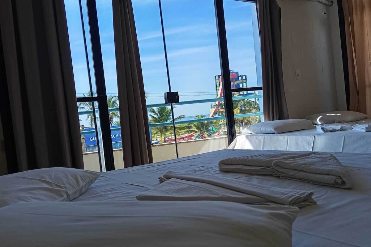 A hotel room with a bed, a window with curtains, a view of a beach, and a pool area.